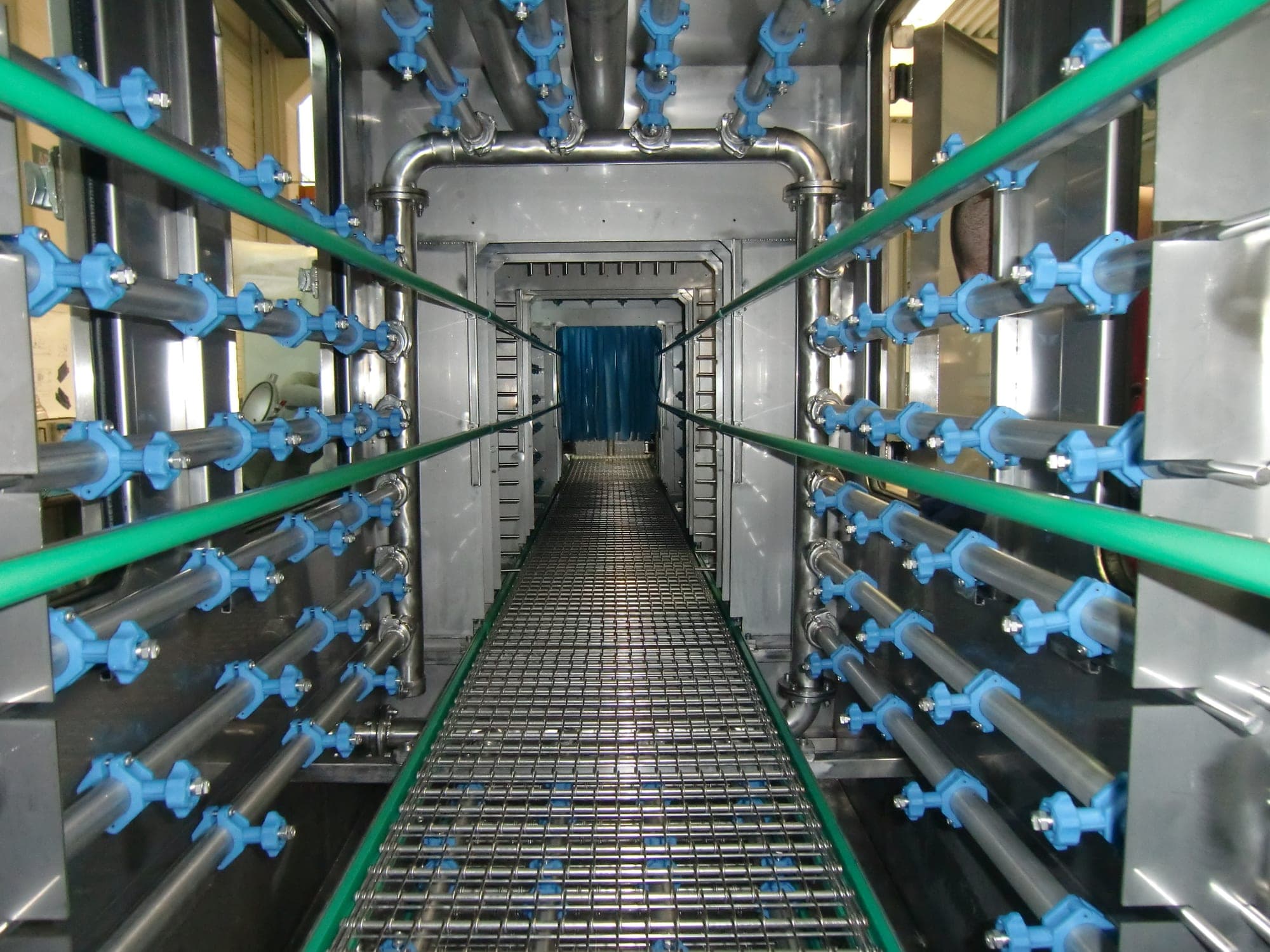 Blick durch das Innere einer Durchlaufreinigungsanlage: beidseitige Reihen von Sprühdüsen über einem Gitterband, das die Bauteile durch die Anlage führt