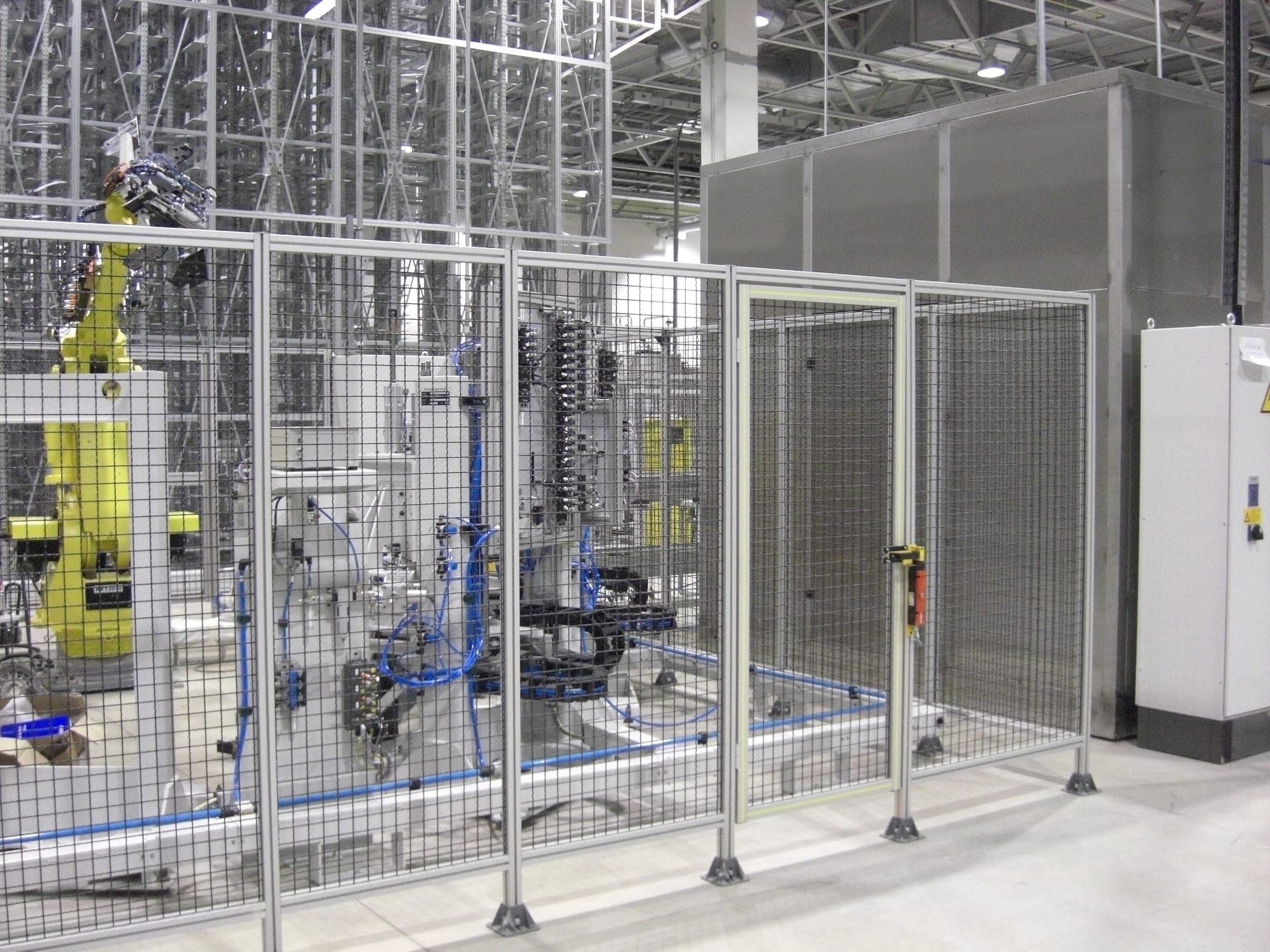 Verkettete Roboterzelle hinter einem Schutzgitter: links ein gelber Knickarmroboter mit Greifer, rechts die Edelstahlkammer einer Reinigungsanlage, dazwischen Handhabungstechnik und Energieketten