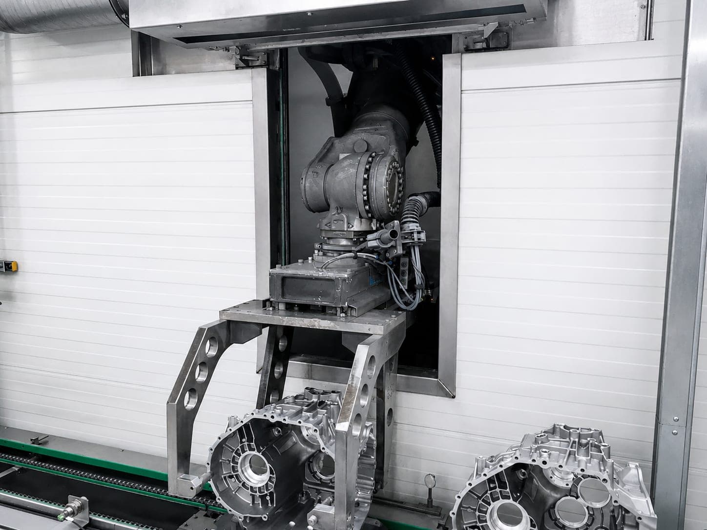 Ausgang einer Entgratzelle: ein Knickarmroboter führt ein Bauteil durch die geöffnete Schleuse nach außen, auf dem Förderband davor stehen zwei Getriebegehäuse aus Aluminiumguss