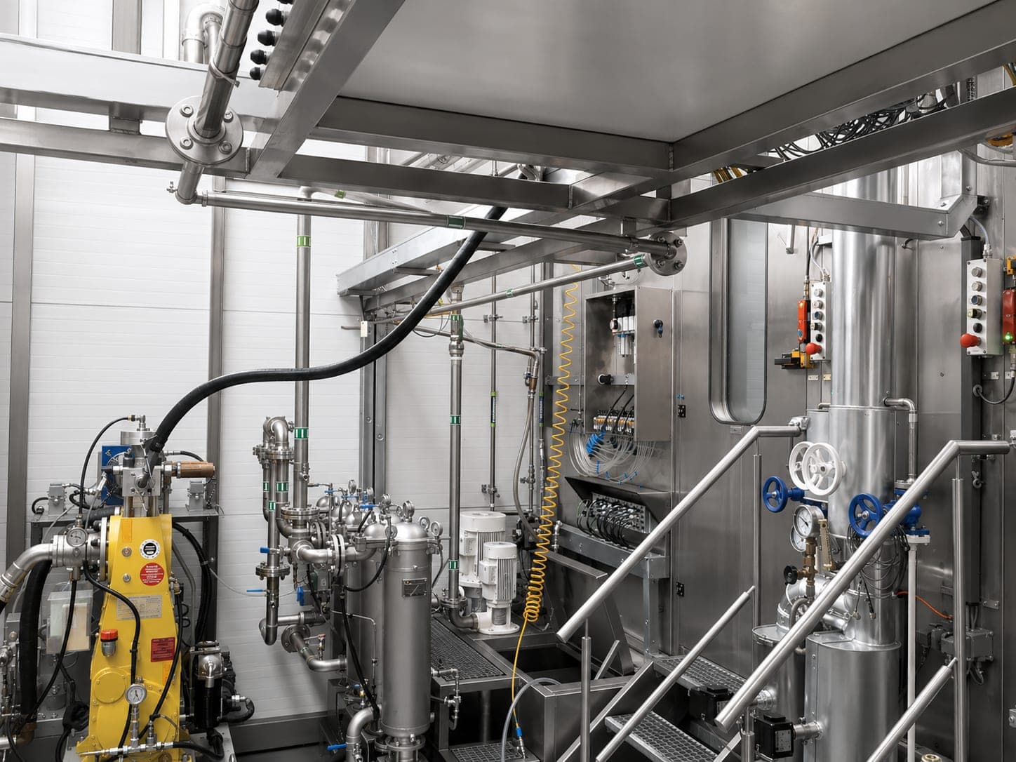 Medien- und Hochdrucktechnik einer Entgratanlage: links die gelb lackierte Hochdruckpumpe mit Manometern, daneben Filtergehäuse, Pumpen und Verrohrung, rechts ein Prozessbehälter mit Handrädern und die Zugangstreppe