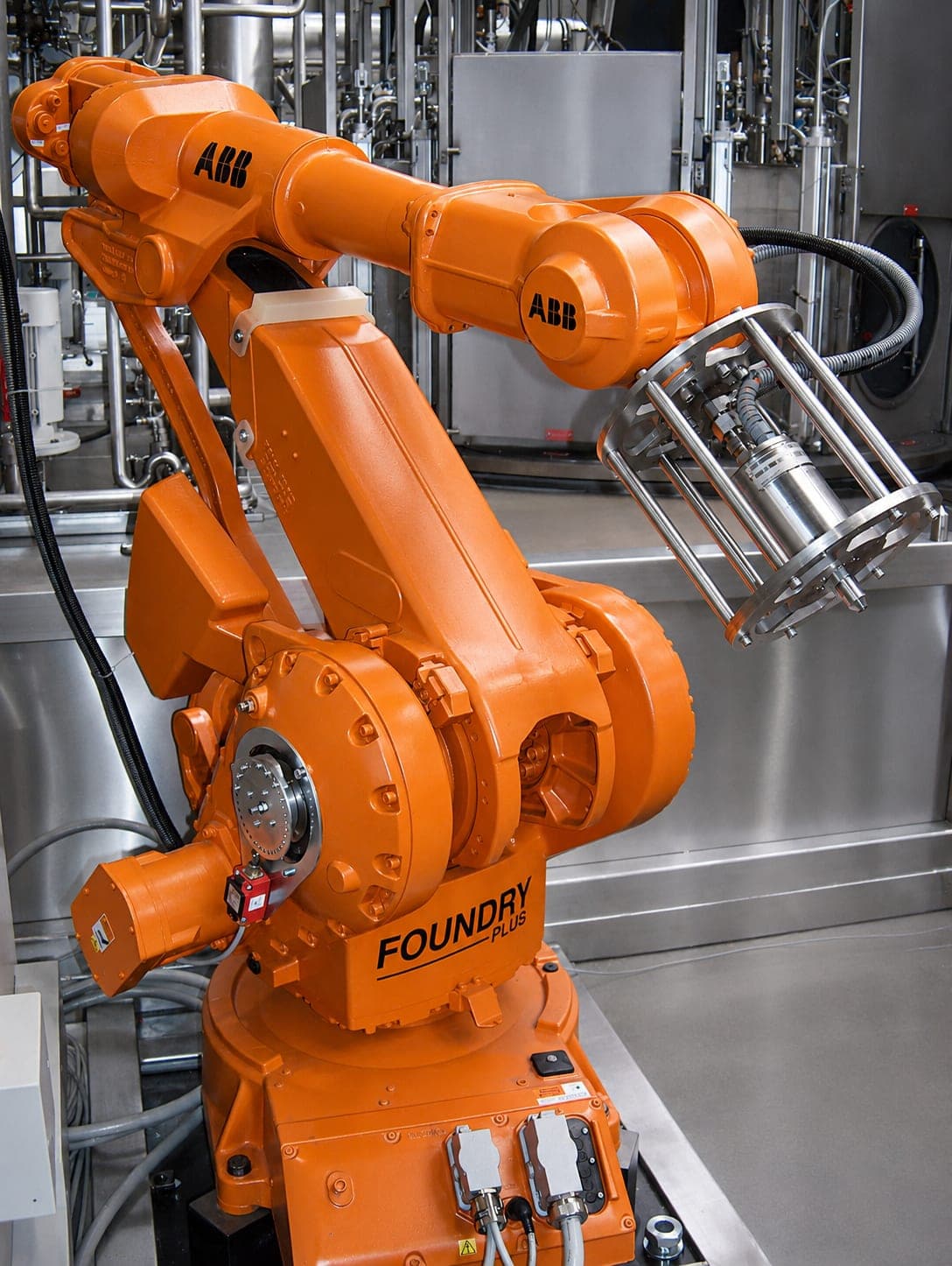 Sechsachsiger Knickarmroboter in einer Reinigungsanlage, am Handgelenk eine käfigartige Greiferaufnahme aus Rundstäben mit zentralem Spannzylinder; dahinter die Verrohrung und ein Prozessbehälter