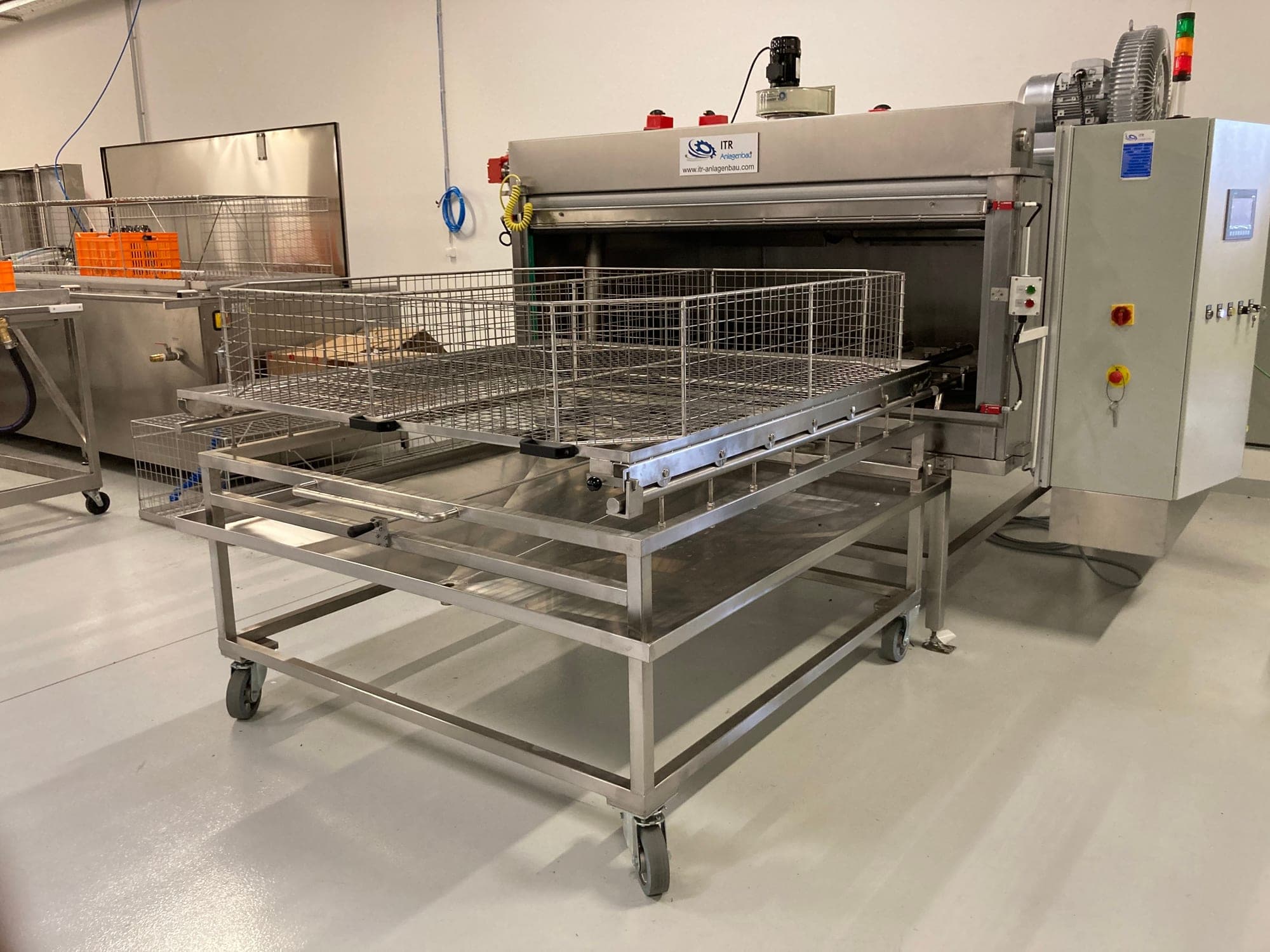 Kompakte Durchlaufreinigungsanlage mit geöffneter Beladeöffnung, davor ein fahrbarer Beladewagen mit Gitterkorb; rechts der Schaltschrank mit Bedienfeld, darüber Seitenkanalverdichter und Signalsäule