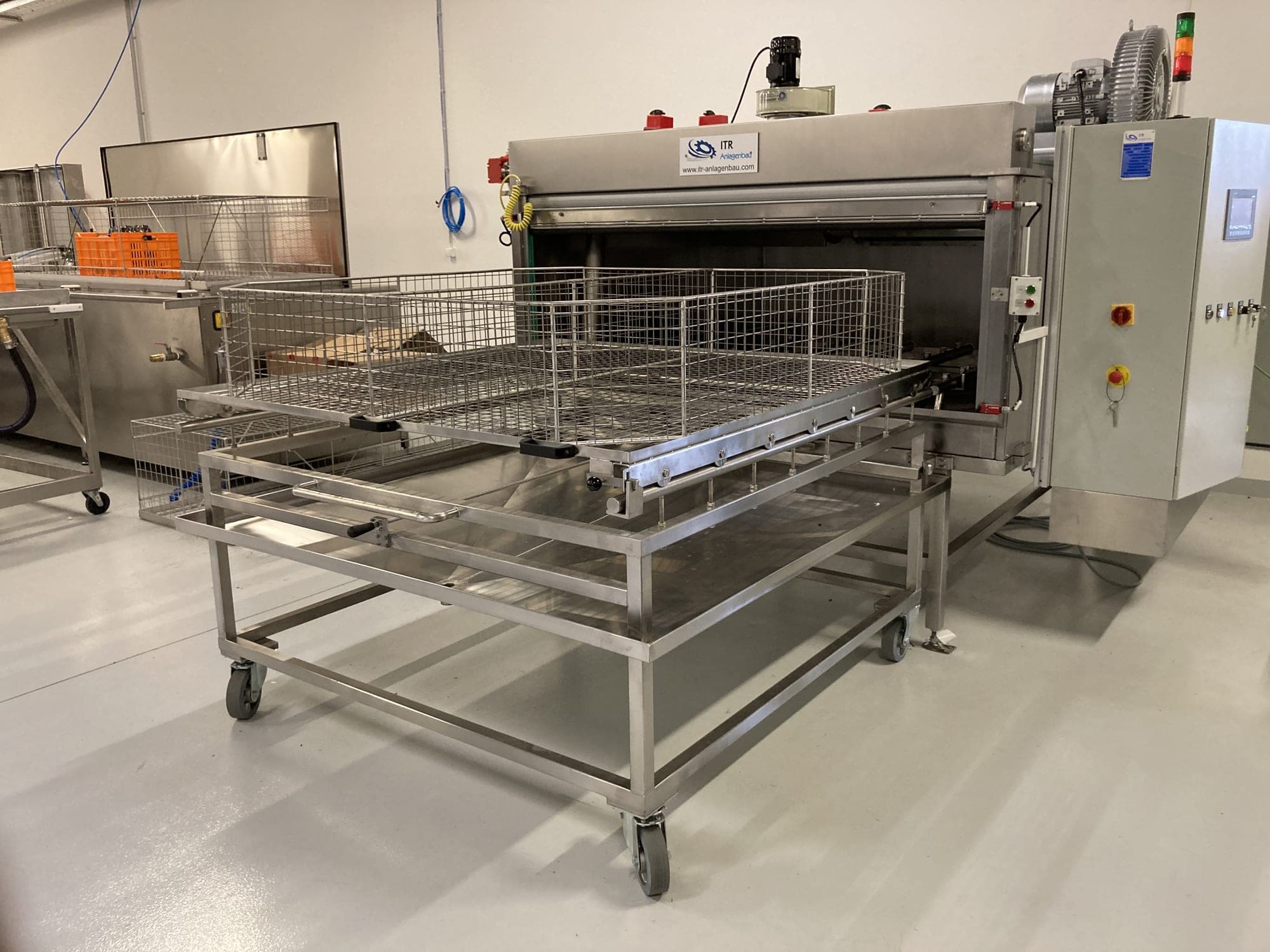 Kammerreinigungsanlage mit geöffneter Fronttür; davor ein Gitterkorb auf einem fahrbaren Beladewagen, rechts daneben der Schaltschrank mit Bedienfeld und Signalsäule