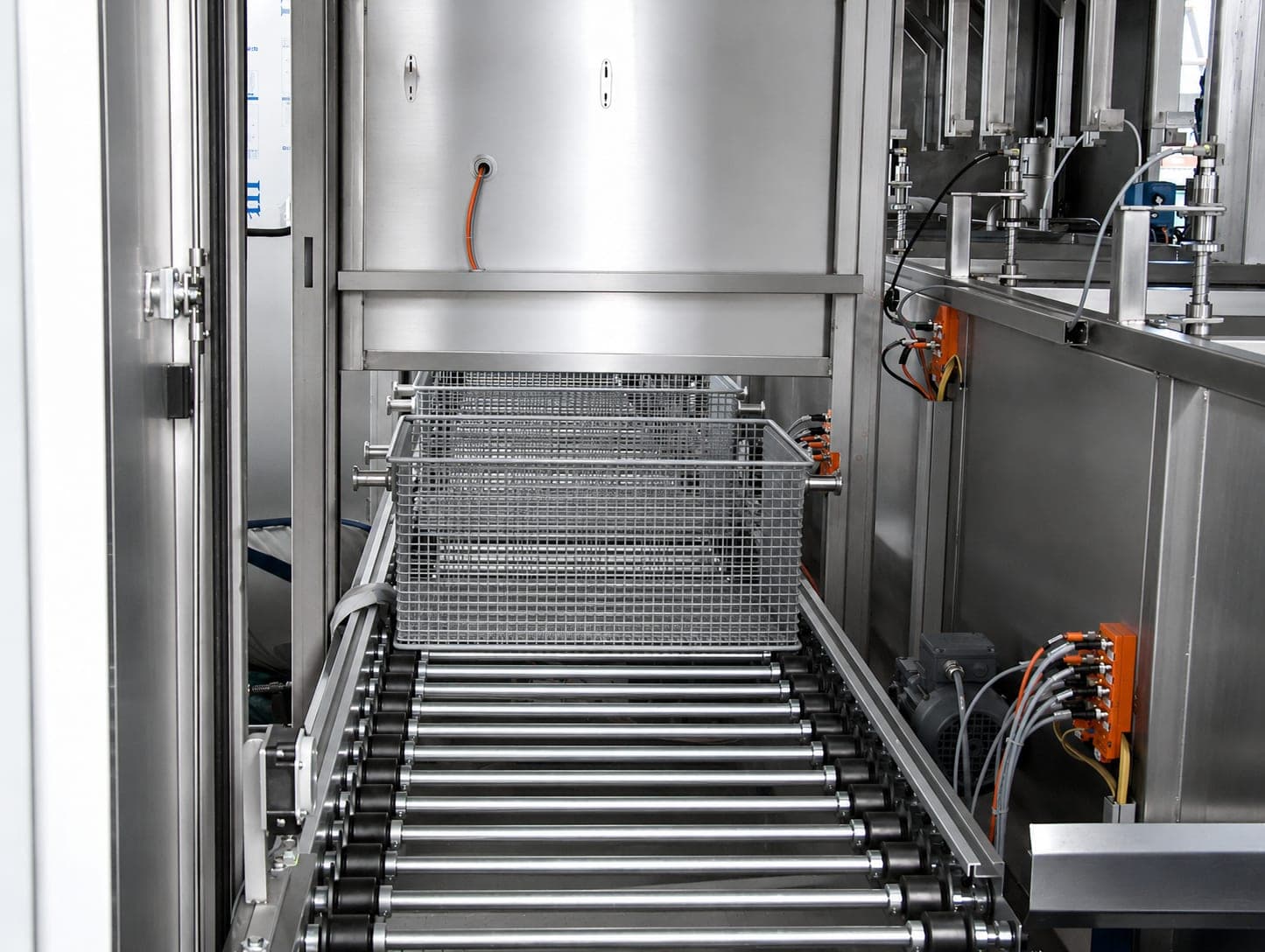 Gitterkorb auf einer angetriebenen Rollenbahn vor der geöffneten Kammer einer Reinigungsanlage, rechts die Antriebe und orange Klemmenkästen der Beladestation