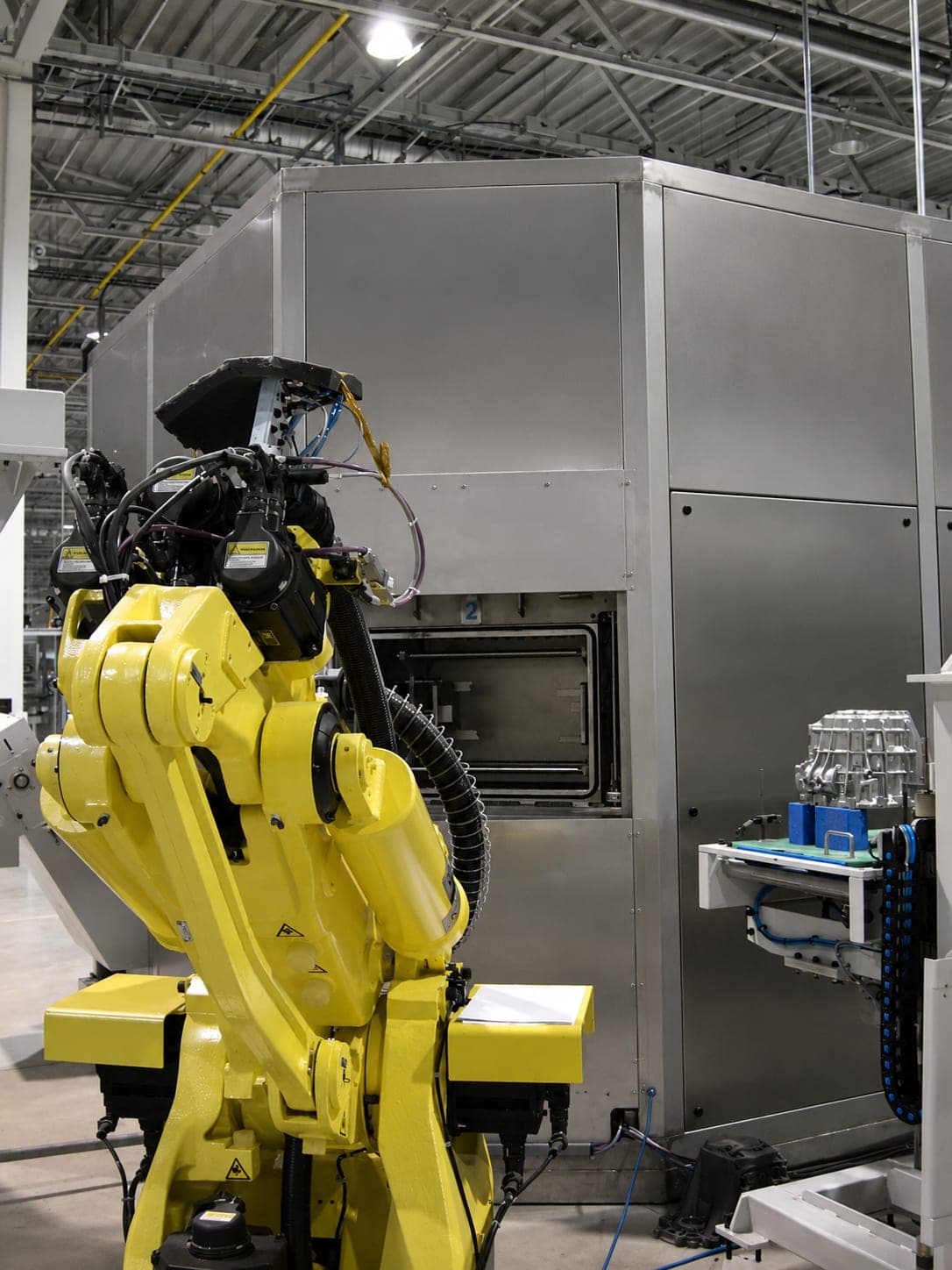 Gelber Knickarmroboter mit Greifer vor der geöffneten Beladeklappe einer eingehausten Reinigungsanlage, rechts daneben ein Gussbauteil auf der Ablage der Zuführung