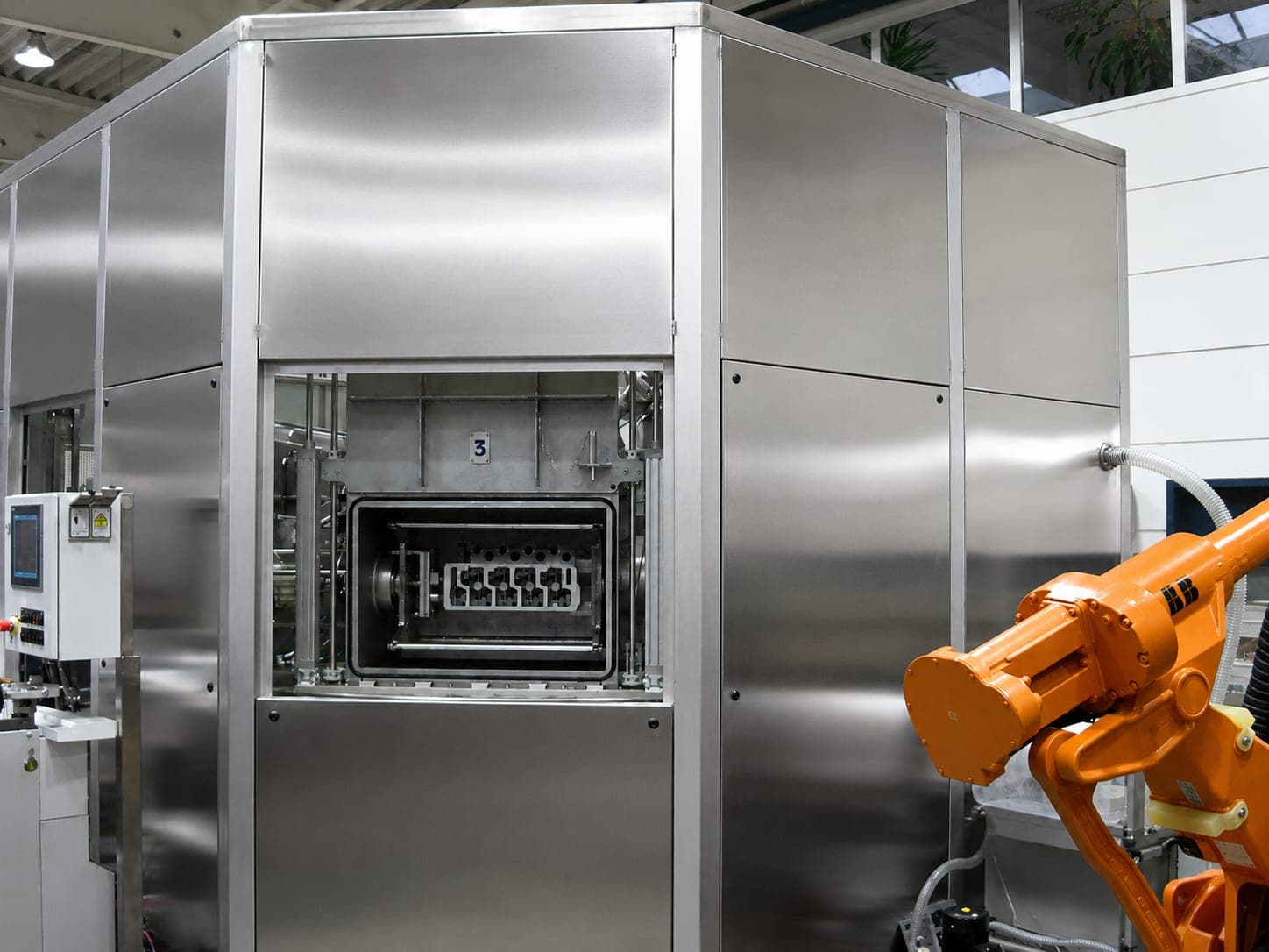 Eingehauste Reinigungsanlage mit geöffneter Kammer: im Innenraum sitzt ein Zylinderkopf auf der drehbaren Werkstückaufnahme, rechts greift ein oranger Knickarmroboter in die Zelle, links das Bedienpanel