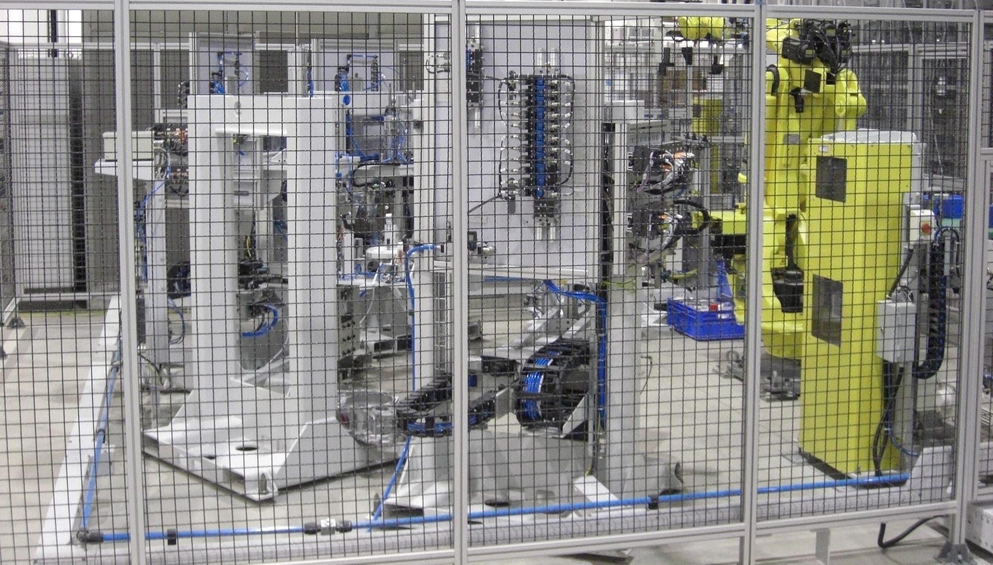 Roboterzelle hinter einem Schutzgitter: rechts ein Knickarmroboter, dahinter Werkstückaufnahmen, Schlauchpakete und Energieketten